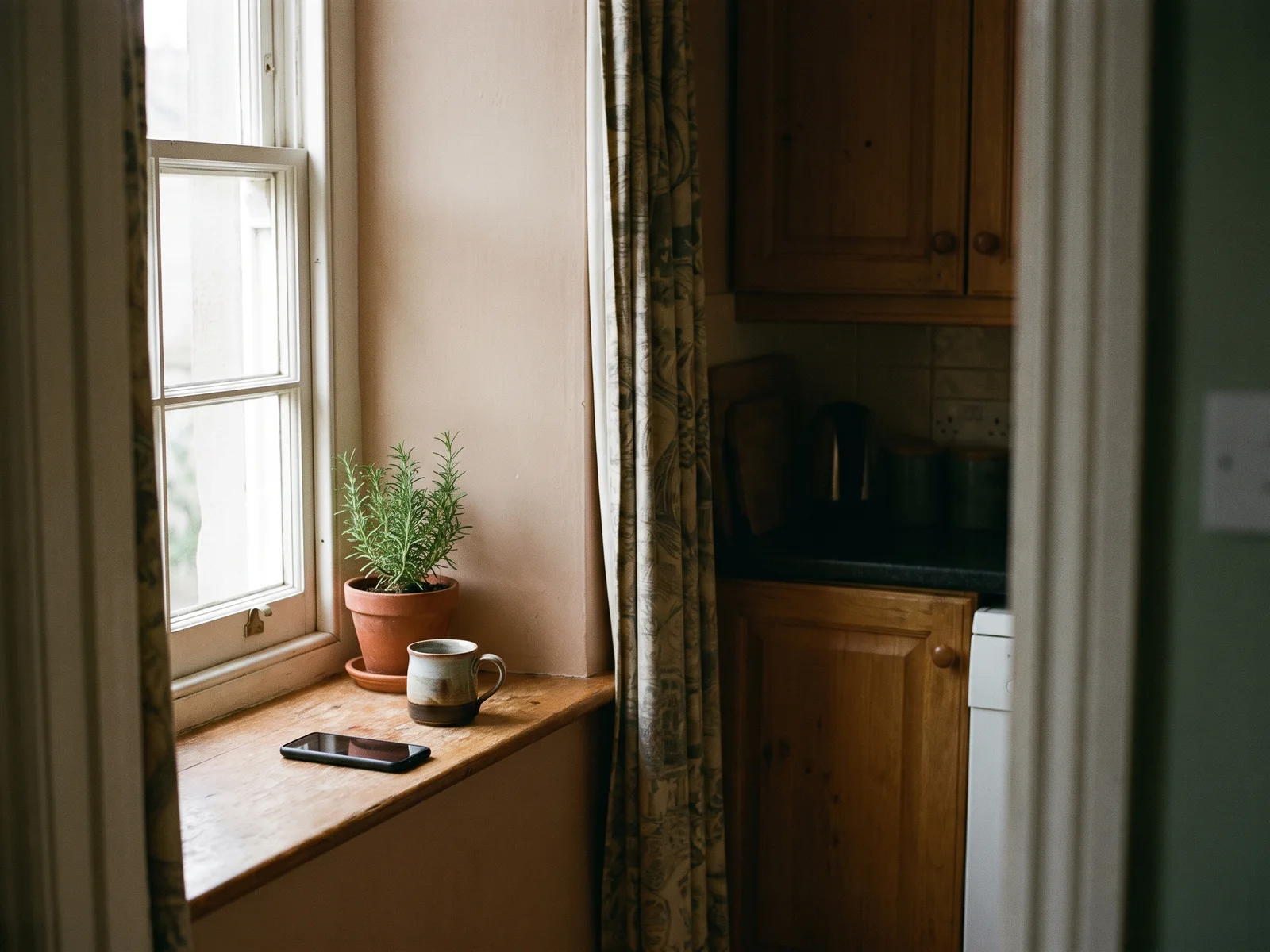 A phone lies face down on a kitchen windowsill beside a mug and a small potted herb in soft side light.