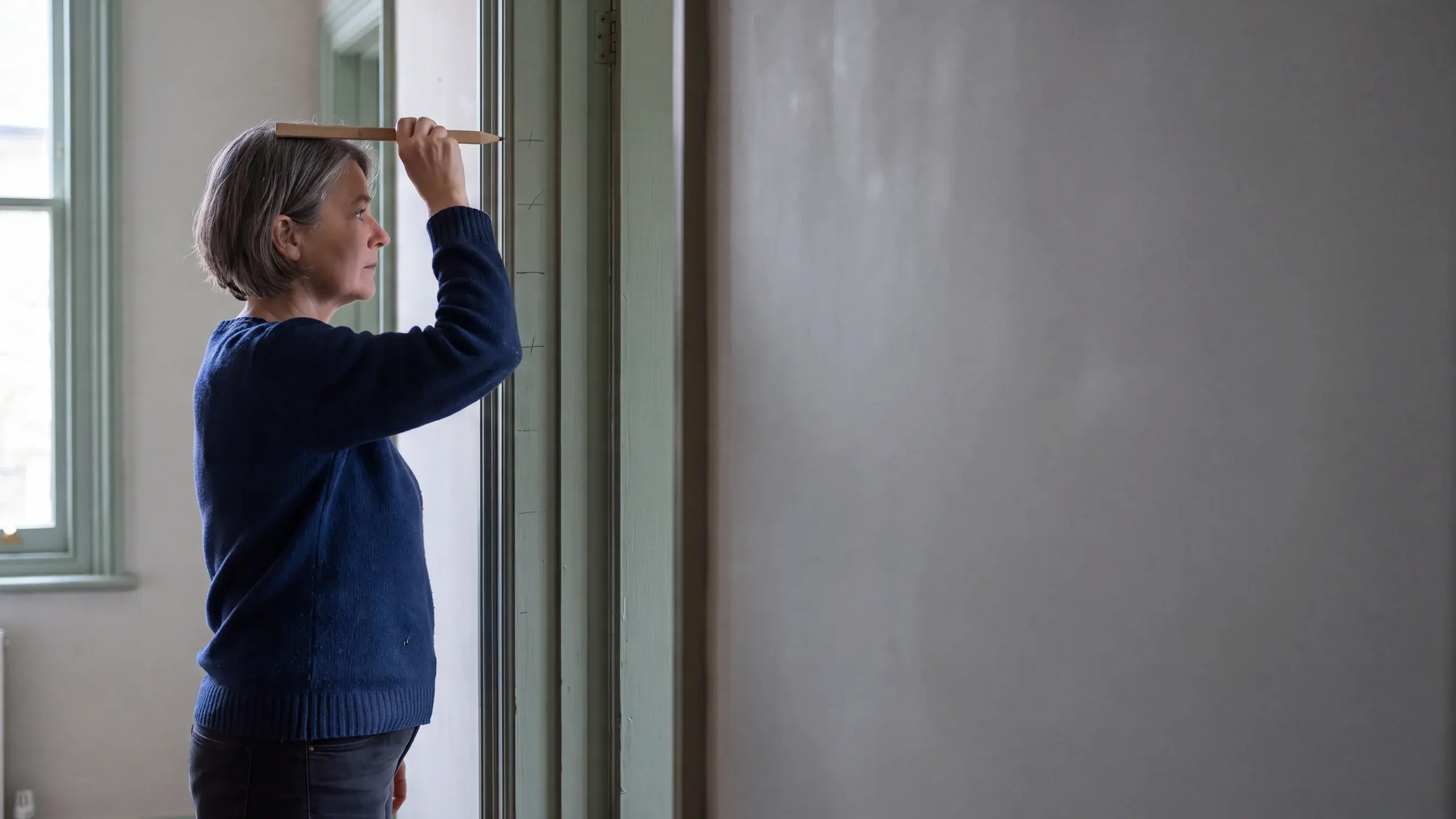 A woman in her fifties stands in profile against a green doorframe, holding a pencil level on top of her head to mark her height.